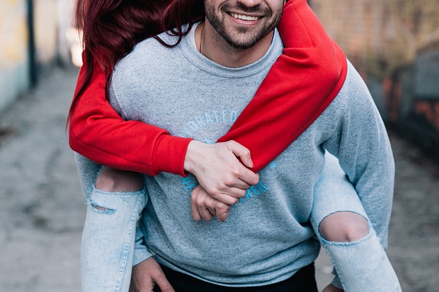 A red-haired woman wearing a red jumper and ripped jeans getting a romantic piggy-back from a smiling man with a grey jumper